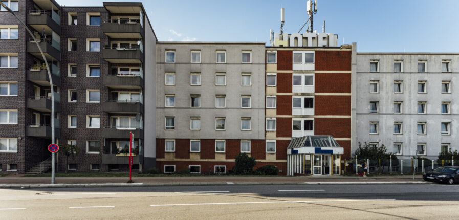 Rechts ist ein grauses Haus mit vielen Fenstern zu sehen. Auf dem Dach steht in großen Buchstaben: Hotel. Links daneben: Ein Wohnhaus.